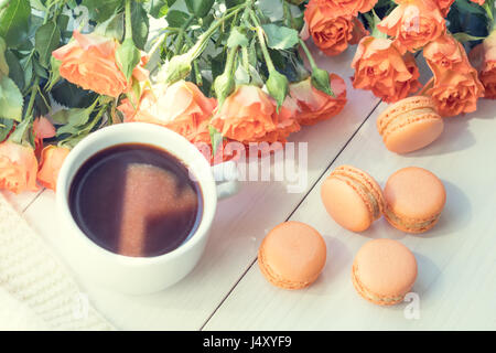 Orange mango or citrous macaroons, cup of coffee and fresh little roses on light wooden background. Sunlight. Coloring and processing photo with light Stock Photo