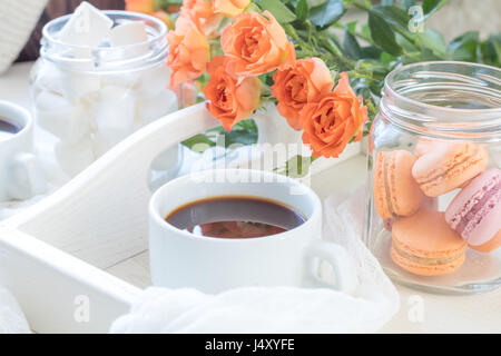 Orange mango macaroons, cup of coffee, marshmallow and fresh little roses on light wooden background. Sunlight. Coloring and processing photo with lig Stock Photo