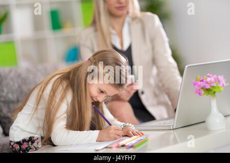 good girl is playing with colored crayons while mom work on laptop in background Stock Photo