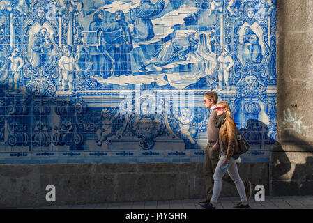 Portugal tiles people, a young couple walk past a wall of blue azulejos tiles on the side of the Capela das Almas church in Bolhao, Porto, Portugal. Stock Photo