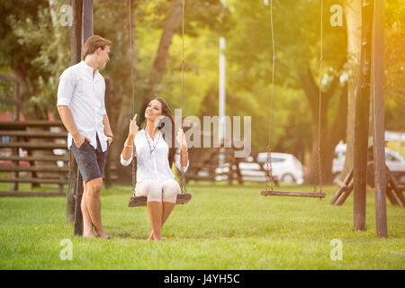 Attractive couple in park on swing, girl sitting on swing man standing next her Stock Photo