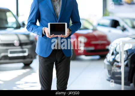 car seller holding empty pc tablet and standing in car in showroom Stock Photo