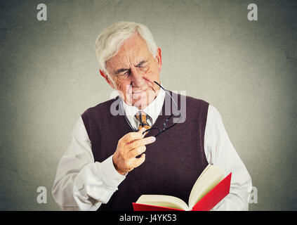 Closeup portrait senior elderly man holding book, glasses having eyesight problems unable to read isolated gray wall background. Human emotion facial  Stock Photo