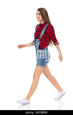 Woman in red lumberjack shirt, jeans dungarees shorts and white sneakers walking and looking away. Side view. Full length studio shot isolated on whit Stock Photo