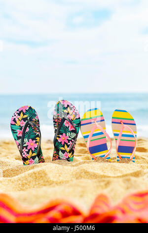 two different pair of colorful flip-flops, one of them flower-patterned and the other striped, stuck on the sand of a quiet beach Stock Photo