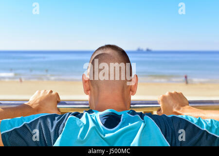 closeup of a young caucasian sportsman in sport clothes seen from behind doing incline push-ups in a metal bar outdoors, next to the sea Stock Photo