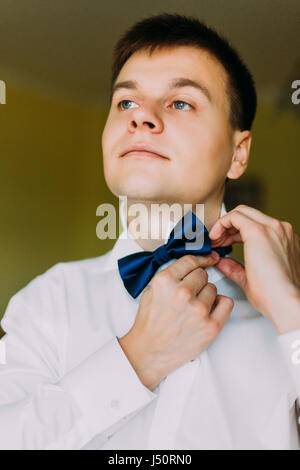 Ellegant handsome groom buttoning his bow tie in a hotel room Stock Photo