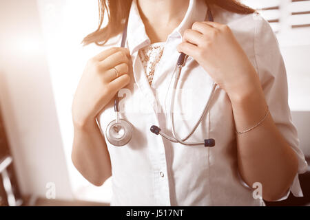 A young female doctor in a white smock holding a stethoscope in arms on neck. Blurred background. Concept of medical help. Stock Photo