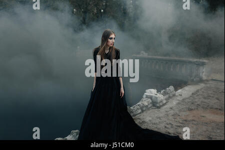 Young attractive Witch in black dress walking on the old bridge in heavy black smoke. Dark magic. Horror fairy tale. Stock Photo