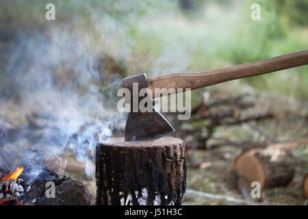 Axe in tree stump and campfire with smoke in summer forest Stock Photo