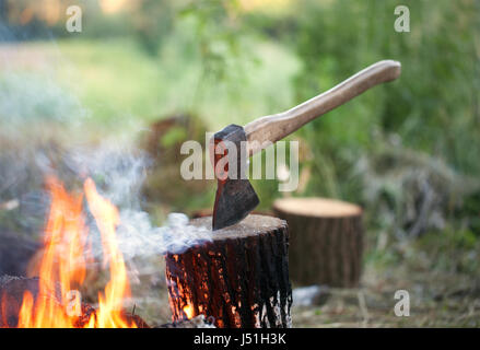 Axe in tree stump and campfire with smoke in summer forest at sun evening Stock Photo