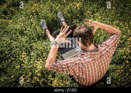Young man sitting on green grass, upset as something is wrong with his laptop Stock Photo