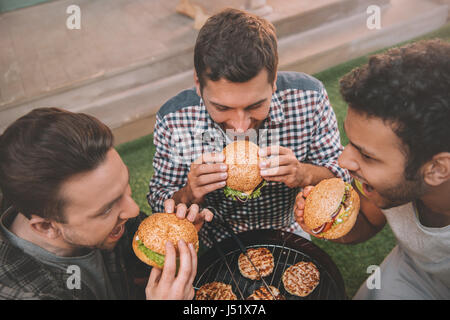 High angle view of three young men eating fresh hamburgers above grill Stock Photo