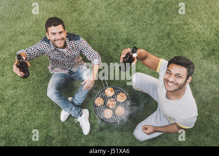 Elevated view of happy young men sitting on grass with beer bottles and making barbecue Stock Photo
