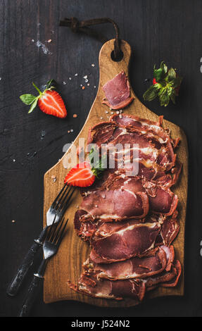 Turkish pastirma with strawberry. Highly seasoned, air-dried cured beef meat cut in slices on wooden board over dark background, top view