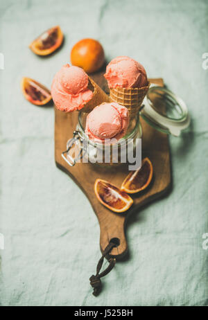 Refreshing summer blood orange ice cream or sorbet scoops in sweet waffle cones in glass jar served with fresh orange slices on rustic wooden board ov Stock Photo