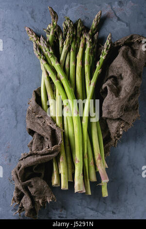 Bundle of young raw uncooked organic green asparagus on sackcloth rag over gray blue metal texture background. Top view. Healthy eating Stock Photo