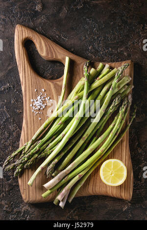 Bundle of young raw uncooked organic green asparagus with sliced lemon and sea salt on wooden chopping board over brown texture background. Top view. Stock Photo