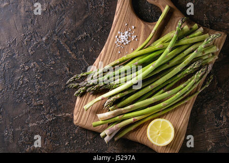 Bundle of young raw uncooked organic green asparagus with sliced lemon and sea salt on wooden chopping board over brown texture background. Top view. Stock Photo