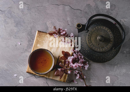 Black iron teapot and traditional ceramic cup of tea on wooden serving board with blossom pink flowers cherry branch over gray texture background. Top Stock Photo