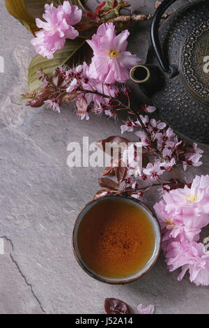 Black iron teapot and traditional ceramic cup of tea with blossom pink flowers cherry branch over gray texture background. Top view with space, Asian Stock Photo