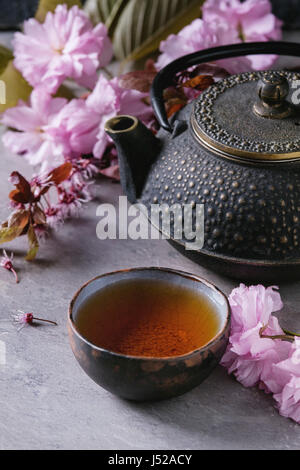 Black iron teapot and traditional ceramic cup of tea with blossom pink flowers cherry branch over gray texture background. Asian style. Stock Photo