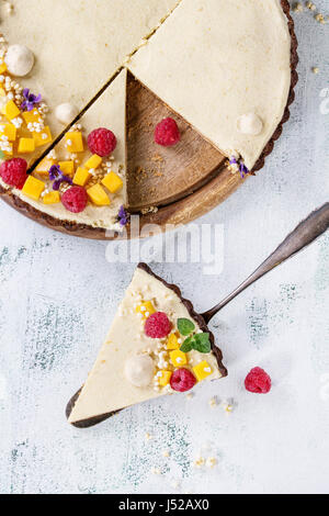 Homemade sliced chocolate tart decorated by mango, raspberries, mint, puffed rice and edible flowers with piece of tart on cake shovel over white text Stock Photo