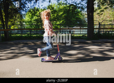 Little blonde girl ride the scooter in the park Stock Photo