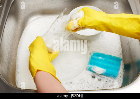 Close up of female hands in yellow protective rubber gloves washing drinking glass with blue cleaning sponge against kitchen sink full of foam and tab Stock Photo