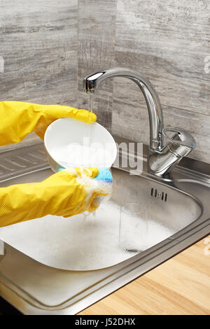 Close up of female hands in yellow protective rubber gloves washing white bowl with blue cleaning sponge against kitchen sink full of foam and tablewa Stock Photo