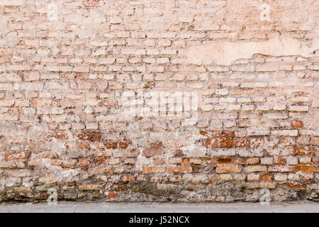 Old weathered vintage brick wall with broken plaster and pavement on the ground. Grungy urban background. Stock Photo