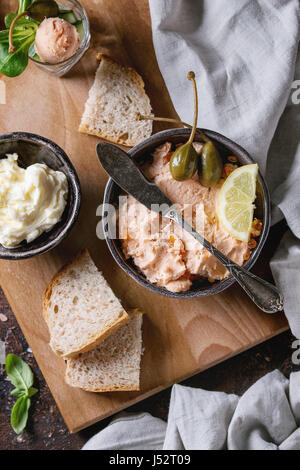 Black bowl of salmon pate with red caviar served with butter, sliced bread, capers, vintage knife, verrines and herbs on wooden serving board, textile Stock Photo