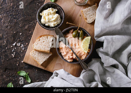 Black bowl of salmon pate with red caviar served with butter, sliced bread, capers, vintage knife and herbs on wooden serving board with textile linen Stock Photo