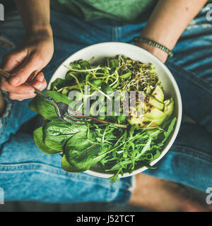 Green vegan breakfast meal in bowl with spinach, arugula, avocado, seeds and sprouts. Girl in jeans holding fork with knees and hands visible, top vie Stock Photo