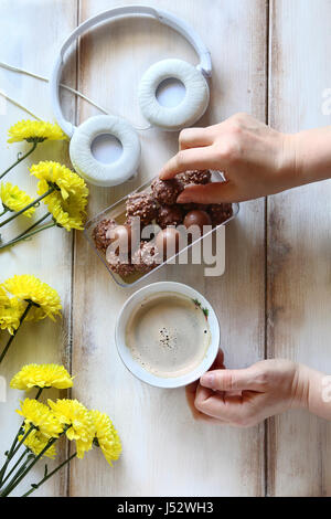 Female hand holding a cup of coffee and serving chocolate candy.Yellow daisy and headphones on the table Stock Photo