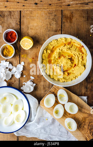 Hummus with hard-boiled egg yolks in a bowl, egg whites in another bowl and on a small wooden cutting board, spices, half lemon and egg shells on a wo Stock Photo