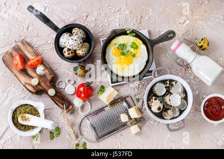 Breakfast with fried quail eggs in iron cast pan, cherry tomatoes, onion, ketchup sauce, seasonings in mortar, cheese grater, egg shell in pot. Beige  Stock Photo