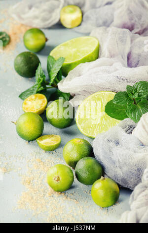 Ingredients for mojito cocktail, whole, sliced lime and mini limes, mint leaves, brown crystal sugar over gray stone texture background with gauze tex Stock Photo