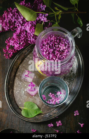 Glass and pitcher of lilac lemonade water with lemon, ice cubes with lilac flowers and lilac branch on vintage iron tray over black wooden table. Dark Stock Photo