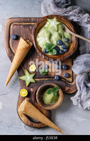 Bowl of Green tea matcha soft elastic ice cream with mint leaves, carambola, lime, blueberries and waffle cones on wooden serving board with wood spoo Stock Photo