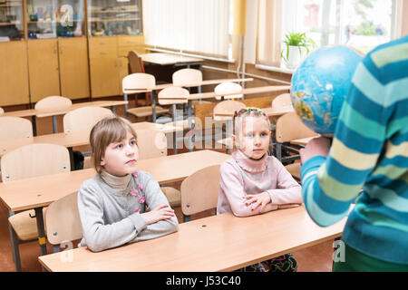 Children sit at school desks in a geography lesson in front of a teacher with a globe Stock Photo