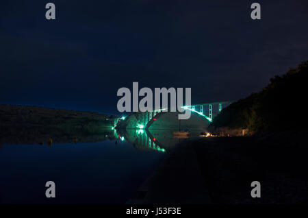 Bridge over sea between an island and mainland illuminated by light with a campfire underneath. Stock Photo