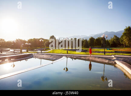 Woman in orange dress near fountain at sunrise in the famous park of Almaty, Kazakhstan Stock Photo