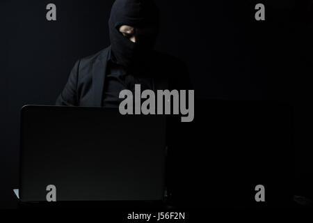 Angry business man as computer hacker in suit stealing data from laptop in front of black background Stock Photo