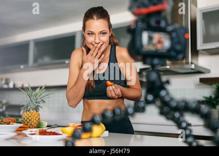 Young lady eating a strawberry at the kitchen table facing the camera. Smiling woman recording content for her vlog in kitchen. Stock Photo