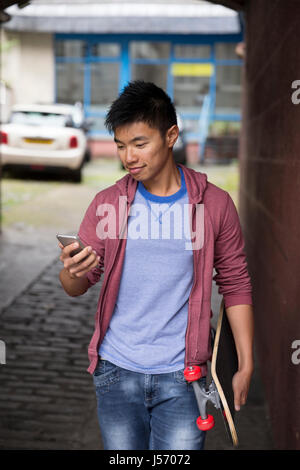 Portrait of a Asian man using his Smart Phone and holding his skateboard. Standing outdoors in city street. Stock Photo