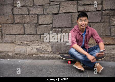 Portrait of a Asian man using his Smart Phone and holding his skateboard. Standing outdoors in city street. Stock Photo