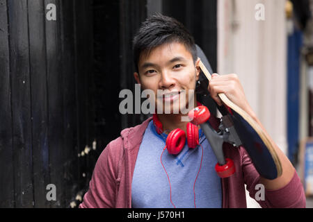 Portrait of a trendy Chinese man with skateboard. Standing outdoors, in front of a wall. Stock Photo