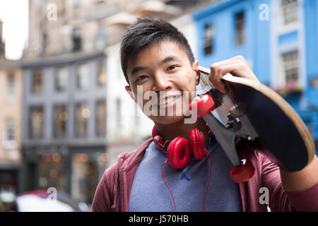 Portrait of a trendy Chinese man with skateboard. Standing outdoors, in front of a wall. Stock Photo
