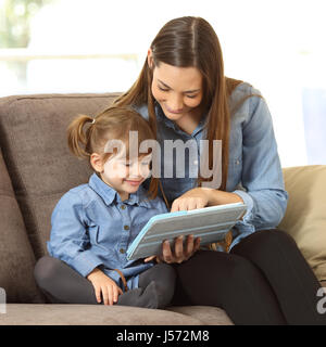 Mother showing media content on line to her two years daughter in a tablet sitting on a couch in the living room in a house interior Stock Photo